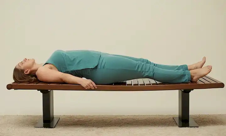 Una persona acostada en un banco inclinado, mirando hacia arriba, con las piernas dobladas a 90 grados, las manos que sostienen la cabeza, los músculos del estómago tensados, la pelvis inclinada, y una curva suave a la espalda, con un fondo neutro